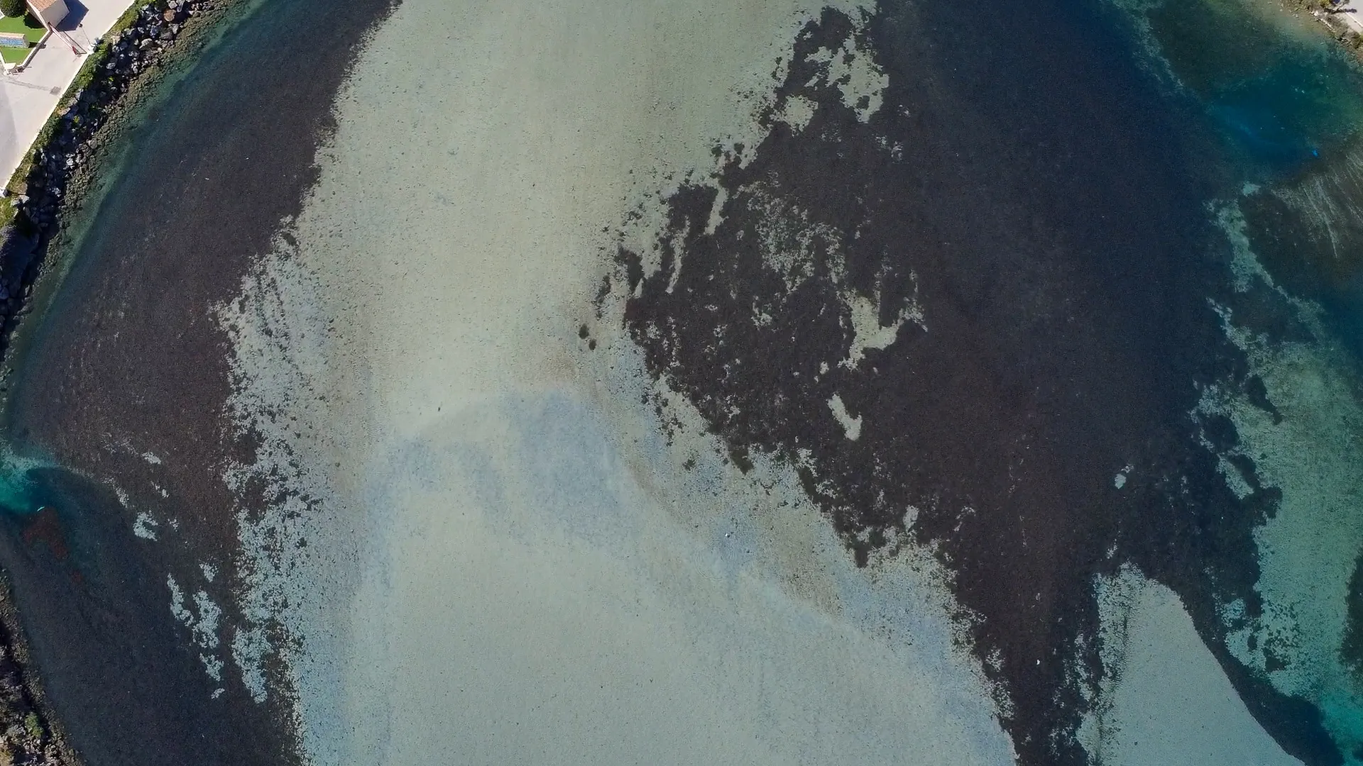 Vista aérea de aguas costeras poco profundas que muestran diversos tonos de azul y marrón, con bordes rocosos y zonas arenosas.